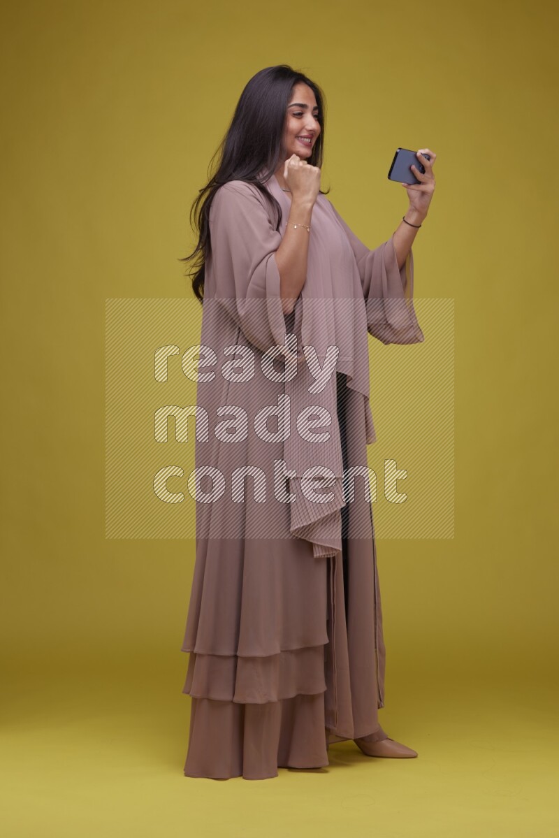 A woman Playing a Game on her smartphone on a Yellow Background wearing Brown Abaya