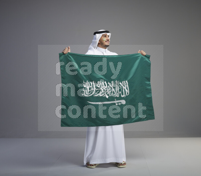 A Saudi man standing wearing thob and white shomag with face painting holding big Saudi flag on gray background
