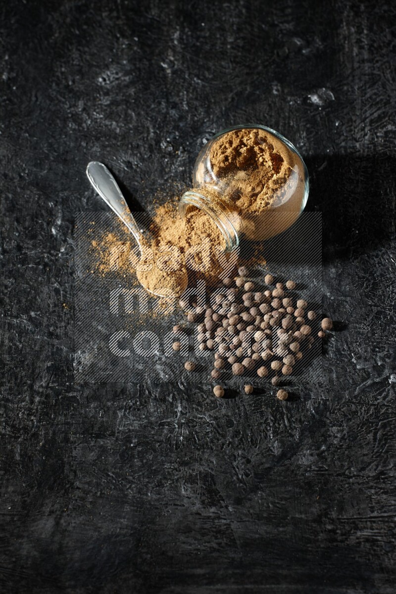 A flipped glass spice jar and metal spoon full of allspice powder and powder spilled out of it with whole balls on a textured black flooring