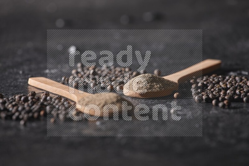 2 wooden spoons full of black pepper powder and black pepper beads spread on a textured black flooring