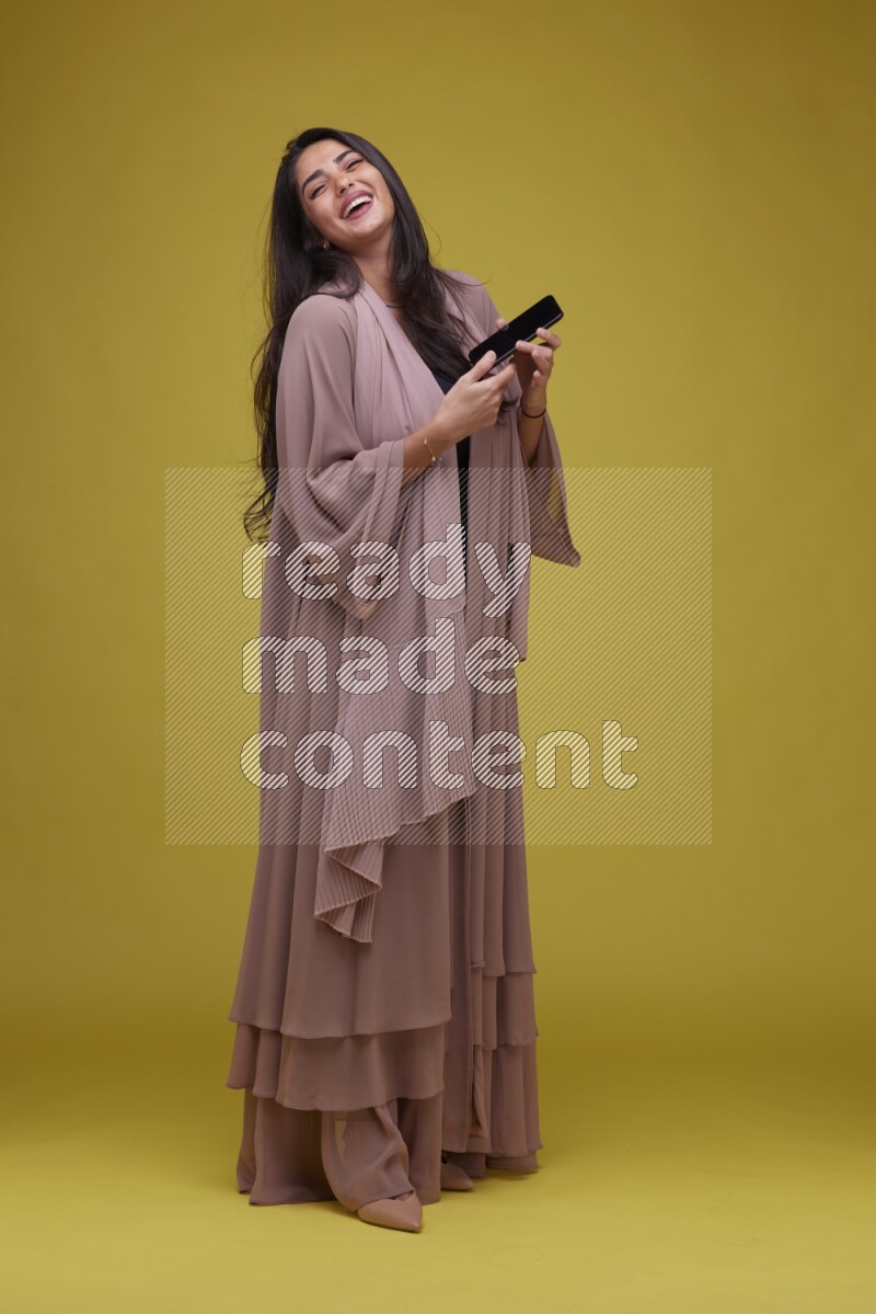 A woman Showing SmartPhone Screen on a Yellow Background wearing Brown Abaya