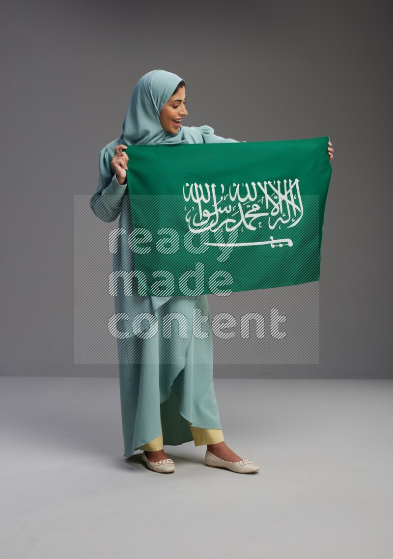 Saudi Woman wearing Abaya standing holding Saudi flag on Gray background