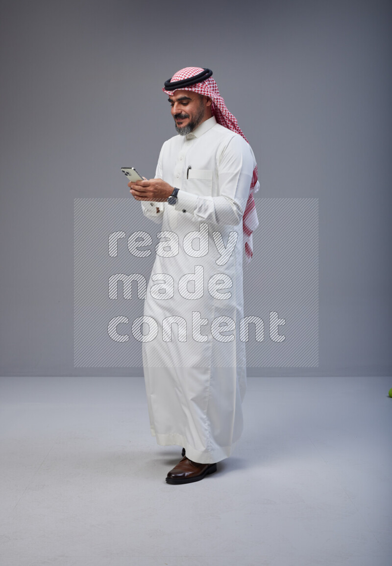 Saudi man Wearing Thob and red Shomag standing texting on phone on Gray background