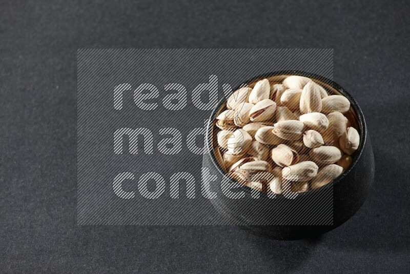A black pottery bowl full of pistachios on a black background in different angles