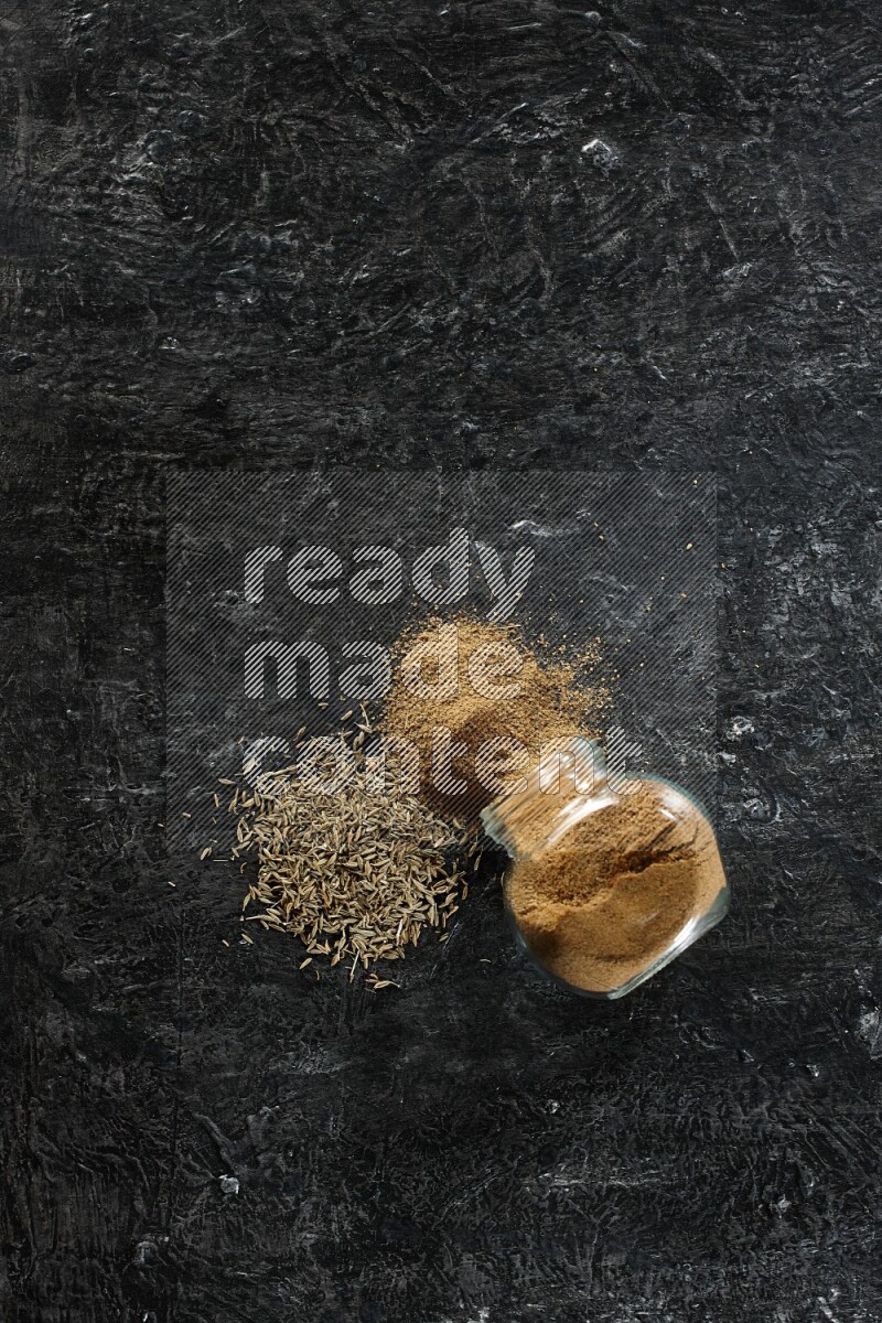 وعاء زجاجي مقلوب ممتلئ ببودرة الكمون مع بودرة متناثرة وحبوب الكمون على خلفية سوداء