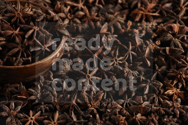 Star Anise in a wooden bowl and surrounded by more anise filling the frame on black flooring