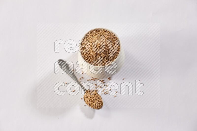 A beige pottery bowl and a metal spoon full of mustard seeds on white flooring