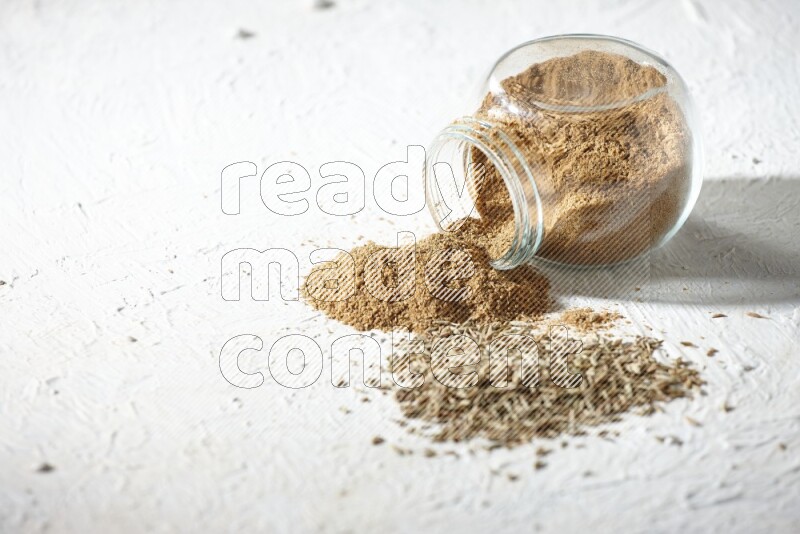 A flipped glass spice jar full of cumin powder and powder spilled out with cumin seeds on textured white flooring