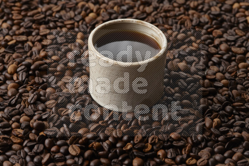 A beige pottery cup of coffee surrounded by roasted coffee beans on beige background