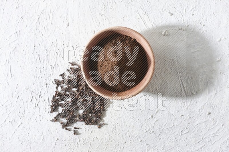 A wooden bowl full of cloves powder with whole cloves beside it on a textured white flooring