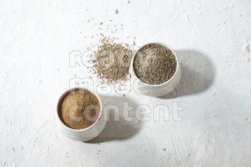 2 beige bowls full of cumin seeds and powder with spilled powder and seeds on textured white flooring