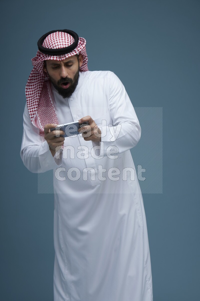 A man Playing a Game on his phone in a blue background wearing Saudi Thob with Shomag