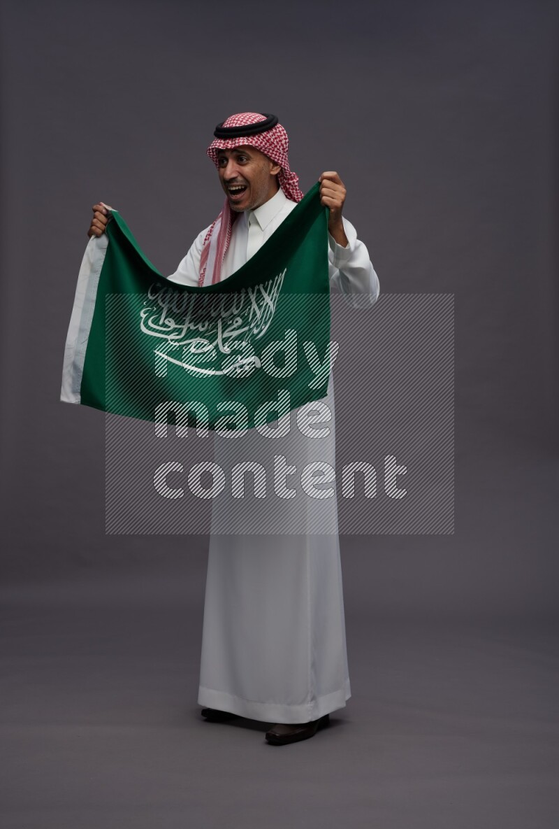Saudi man wearing thob and shomag standing holding Saudi flag on gray background