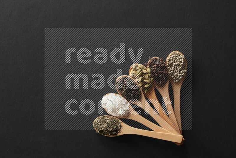 wooden spoons filled with white peppers, cloves, cardamom, salt, black peppers and basil on black flooring