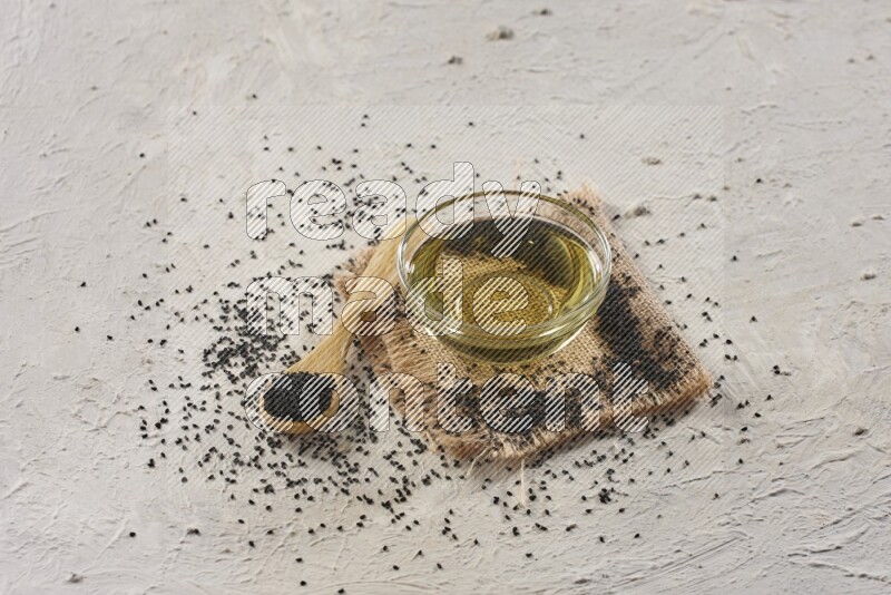 وعاء زجاجي ممتلئ بزيت حبة البركة وملعقة خشبية مليئة ببذور حبة البركة مع بعض البذور المتناثرة علي قطعة من القماش على أرضية بيضاء