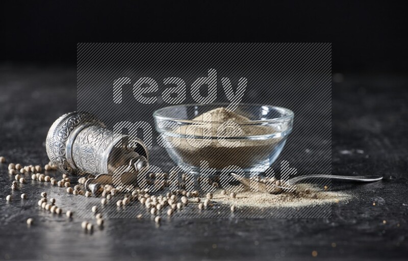 A glass bowl full of white pepper powder with pepper beads, a metal grinder and a metal spoon on textured black flooring