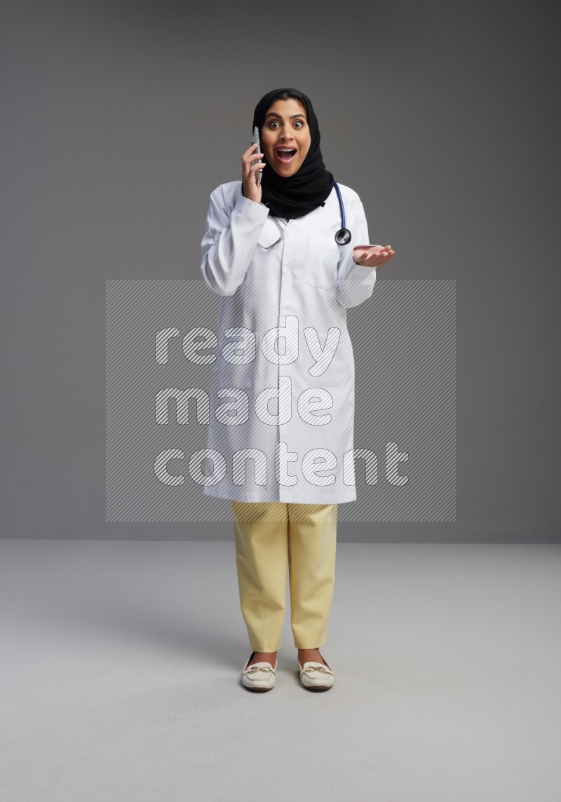 Saudi woman wearing lab coat with stethoscope standing talking on phone on Gray background