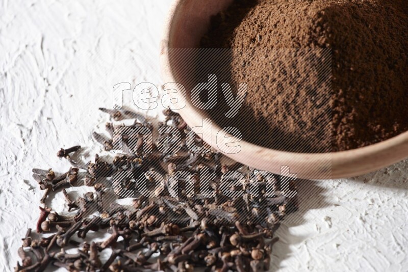 A wooden bowl full of cloves powder with whole cloves beside it on a textured white flooring
