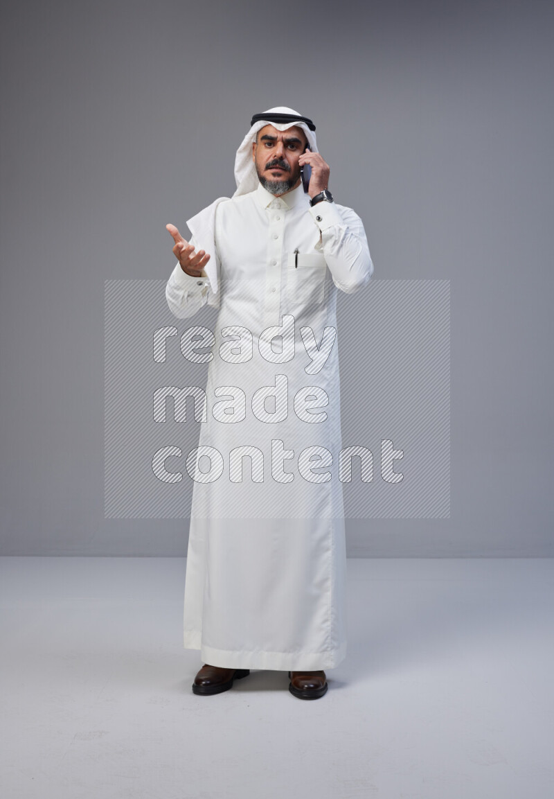 Saudi man Wearing Thob and white Shomag standing talking on phone on Gray background