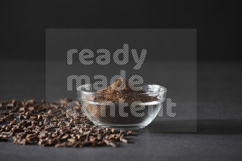 A glass bowl full of cloves powder with cloves grains spread on black flooring
