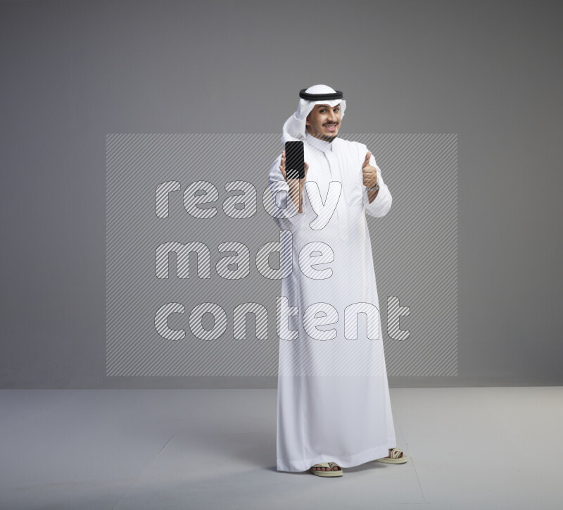 A Saudi man standing wearing thob and white shomag showing phone to camera on gray background