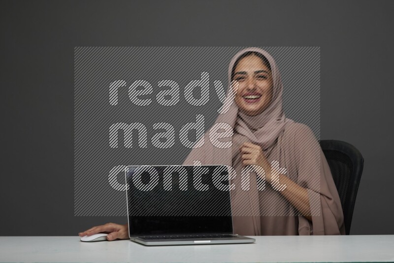 A woman Sitting on her desk  Pointing at her laptop on a Gray Background wearing Brown Abaya with Hijab