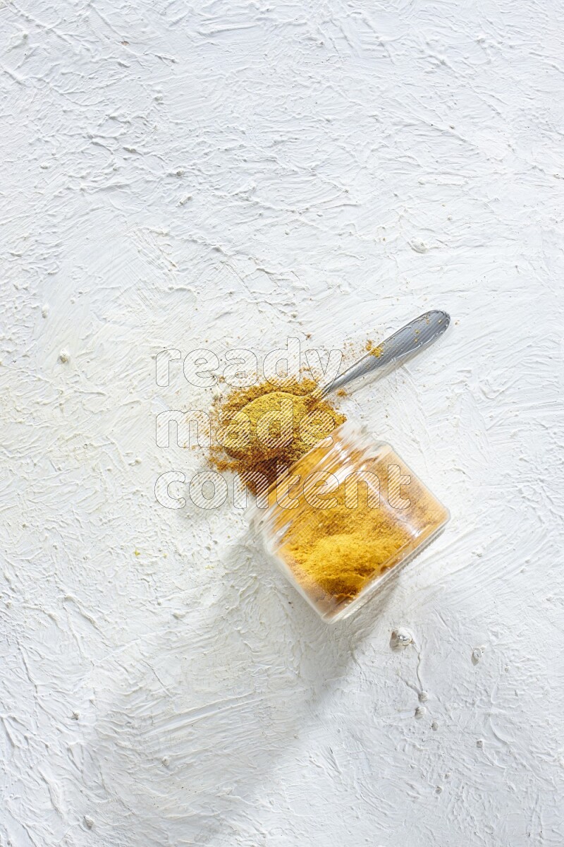 A flipped glass spice jar and a metal spoon full of turmeric powder and powder spilled out of it on textured white flooring