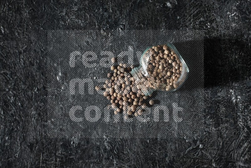 A flipped glass spice jar full of allspice whole balls and the balls spilled out of it on a textured black flooring