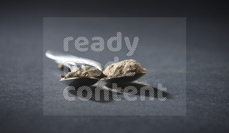 ملعقتان معدنيتان ممتلئتان ببودرة الهيل وحبوب الهيل على خلفية سوداء