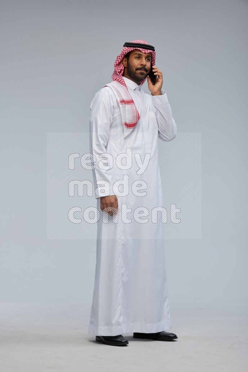Saudi man Wearing Thob and shomag standing talking on phone on Gray background