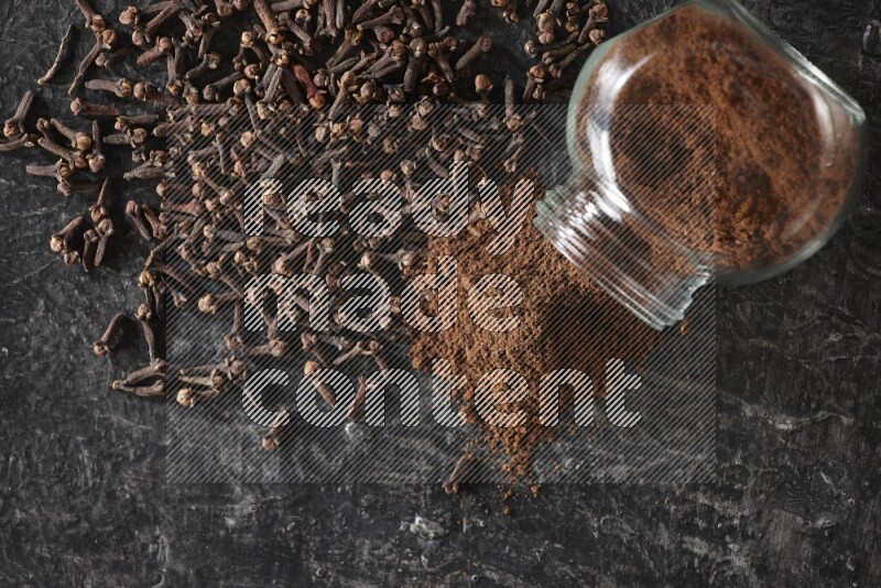 وعاء زجاجي للبهارات مقلوب ممتلئ ببودرة القرنفل والبودرة سقطت منه مع حبوب القرنفل المتناثرة على أرضية سوداء