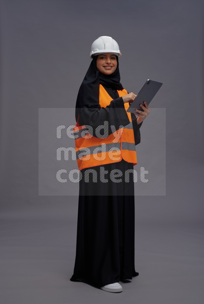 Saudi woman wearing Abaya with engineer vest standing working on tablet on gray background