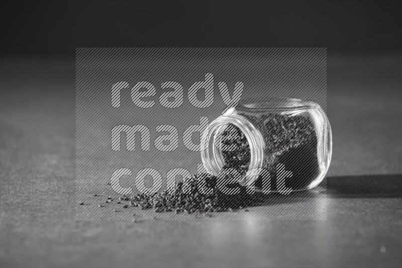 A flipped glass spice jar full of black seeds and the seeds spread out on a black flooring