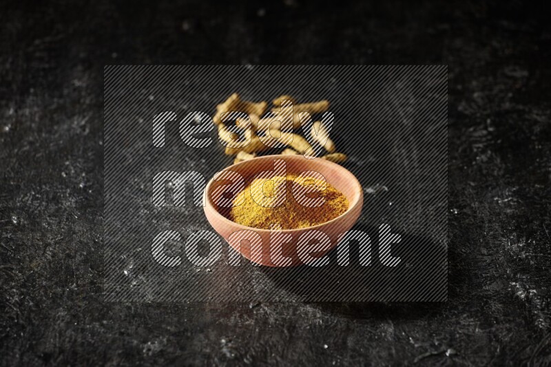 A wooden bowl full of turmeric powder with dried turmeric fingers on textured black flooring