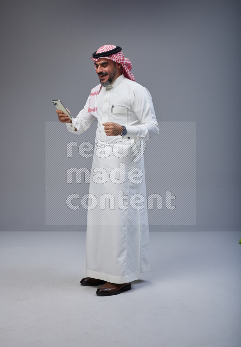 Saudi man Wearing Thob and red Shomag standing texting on phone on Gray background