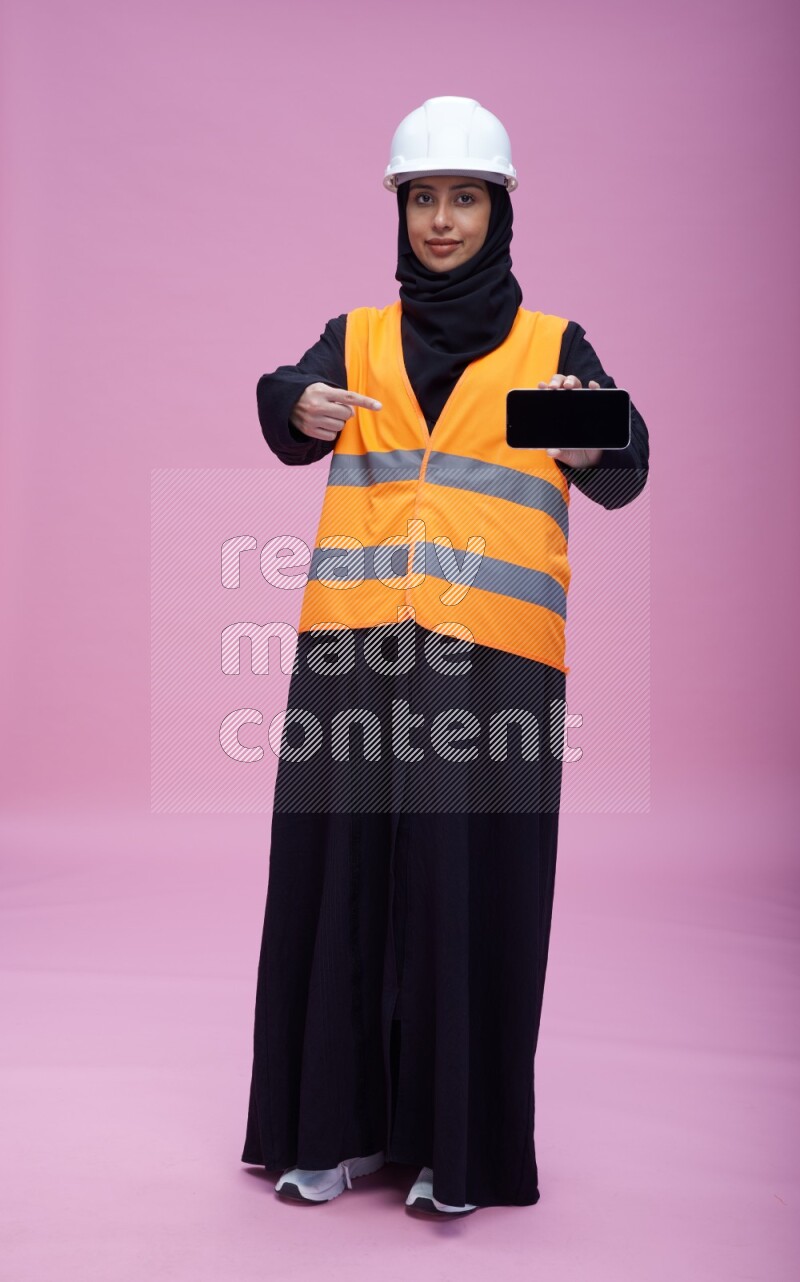 Saudi woman wearing Abaya with engineer vest and helmet standing showing phone to camera on pink background