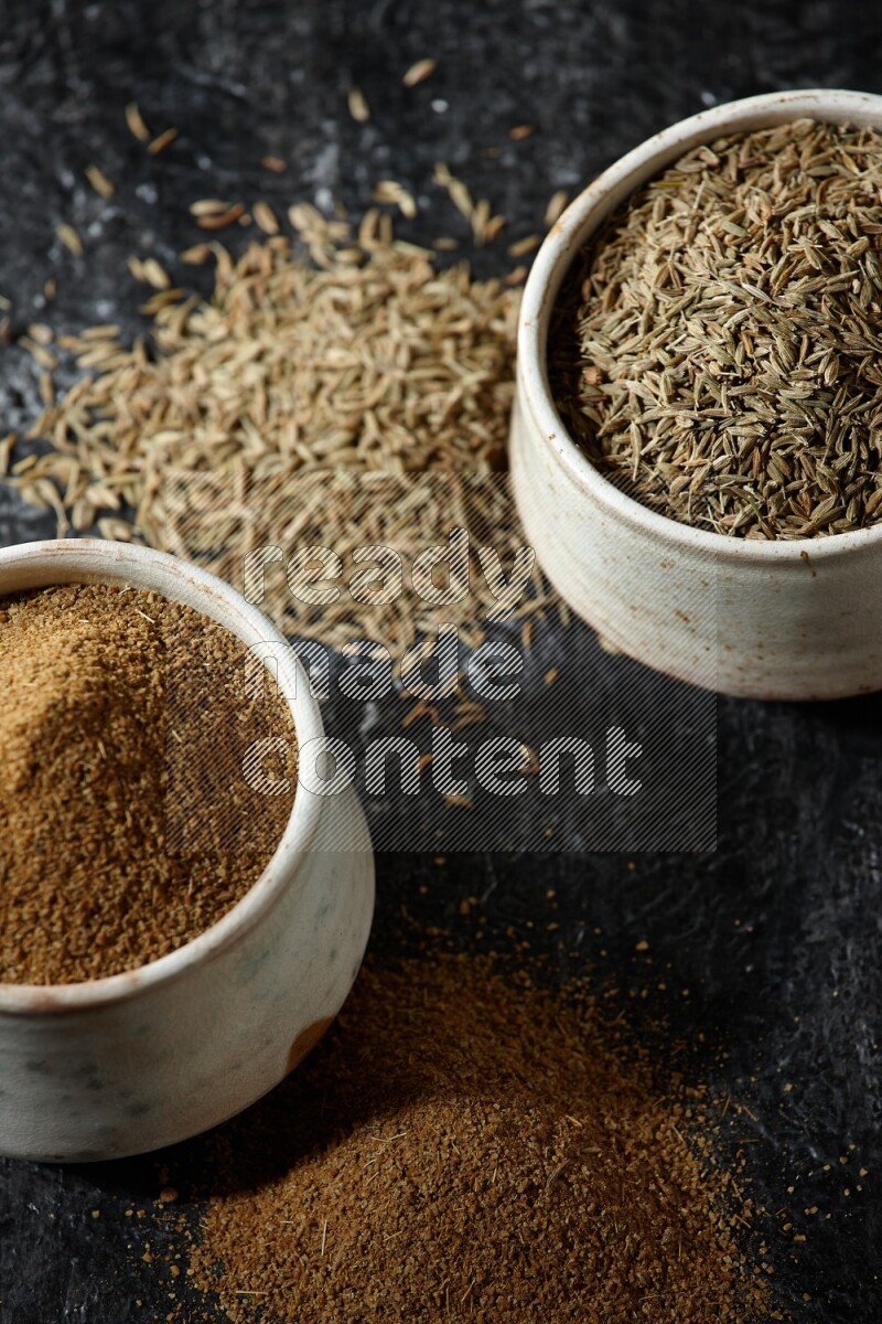 2 beige bowls, one full of cumin powder and the other full of seeds on a textured black flooring