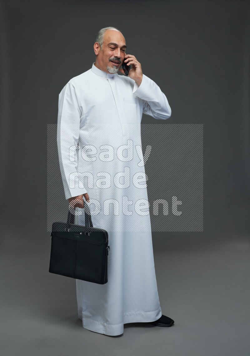 Saudi man without shomag Standing holding bag and talking on phone on gray background