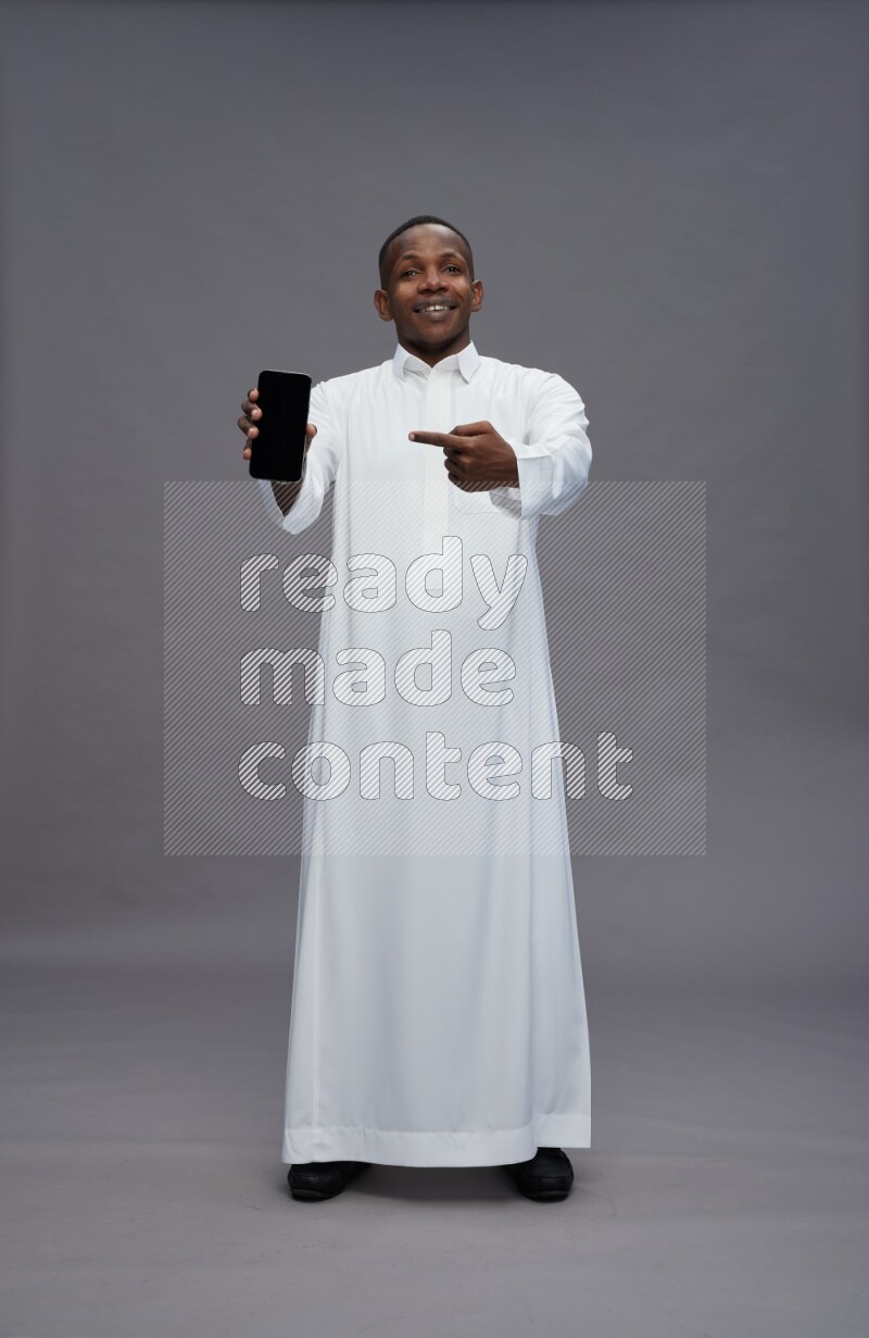 Saudi man wearing thob standing showing phone to camera on gray background