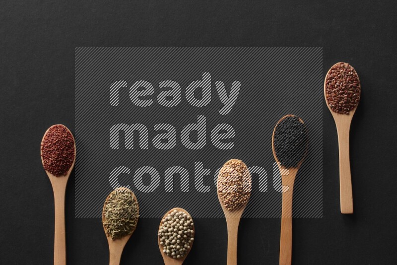 wooden spoons filled with white peppers, mustard seeds, black seeds, garden cress, cumin and flax on black flooring