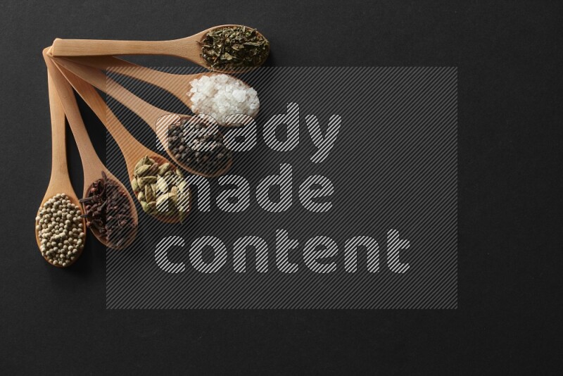 wooden spoons filled with white peppers, cloves, cardamom, salt, black peppers and basil on black flooring