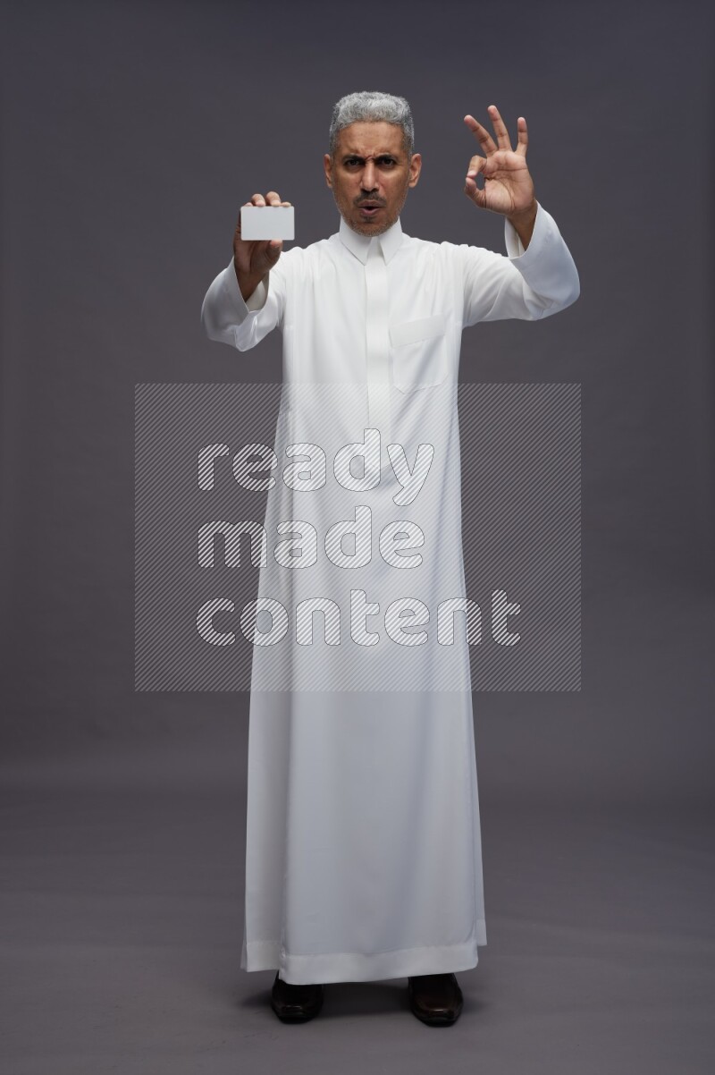 Saudi man wearing thob standing holding ATM card on gray background