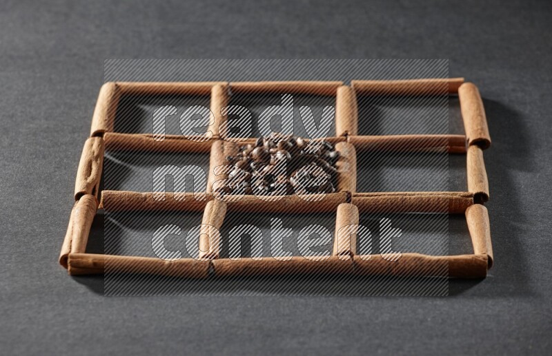 9 squares of cinnamon sticks full of coffee beans in the middle surrounded by nutmeg, dried mint, cloves, dried basil, dried ginger, cinnamon, star anise and cardamom on black flooring
