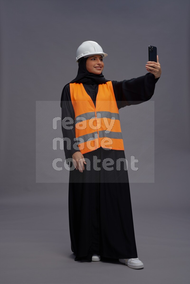 Saudi woman wearing Abaya with engineer vest standing taking selfie on gray background