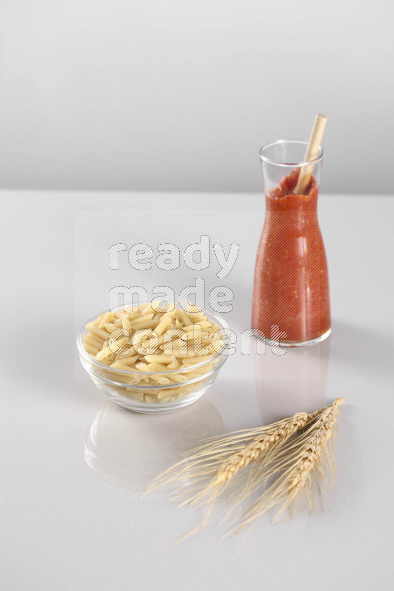Raw pasta with tomatoe pasta with different ingredients such as cherry tomatoes, basil, garlic, bay laurel, cardamom, white pepper, black pepper, red chilis and wheat stalks on light grey background