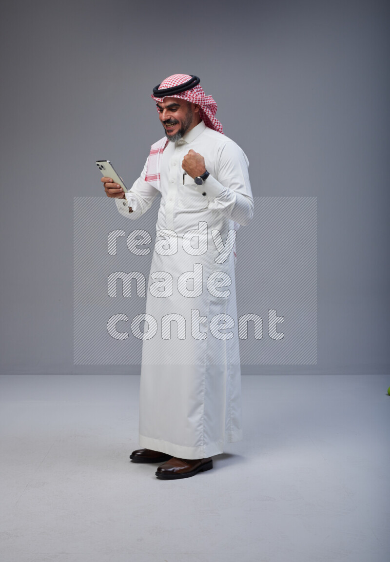 Saudi man Wearing Thob and red Shomag standing texting on phone on Gray background