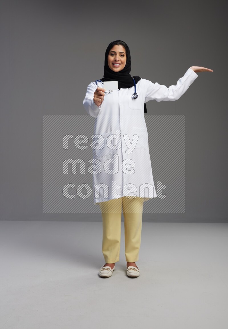 Saudi woman wearing lab coat with stethoscope standing holding ATM card on Gray background