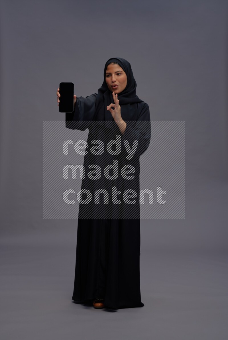 Saudi woman wearing Abaya standing showing phone to camera on gray background