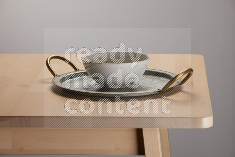 off white bowl with islamic pattern placed on a rounded stainless steel tray with golden handels on the edge of wooden table