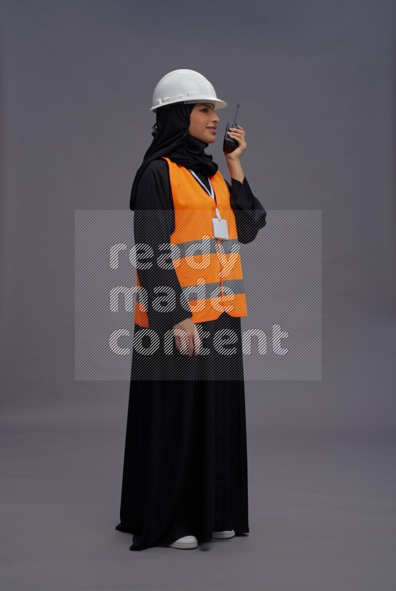 Saudi woman wearing Abaya with engineer vest with neck strap employee badge standing holding walkie-talkie on gray background